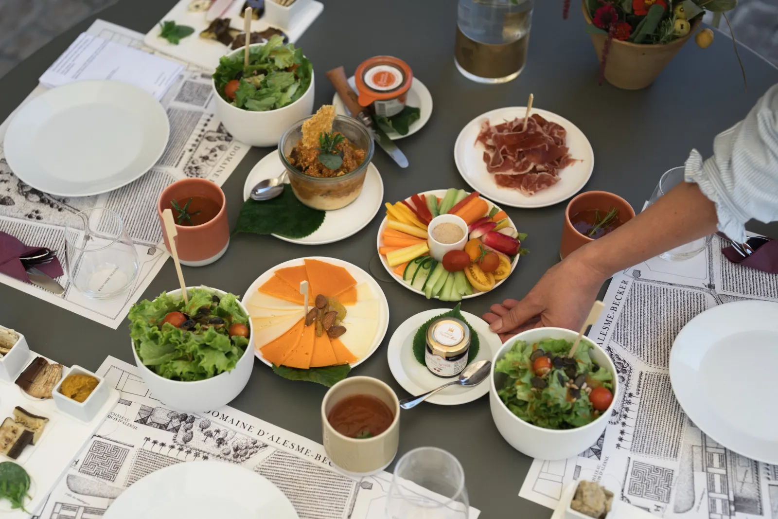 Gourmet tasting table at Château Marquis d'Alesme in Bordeaux, featuring fresh salads, cured meats, a cheese and fruit board, accompanied by wine and water glasses, with newspapers spread on the table.