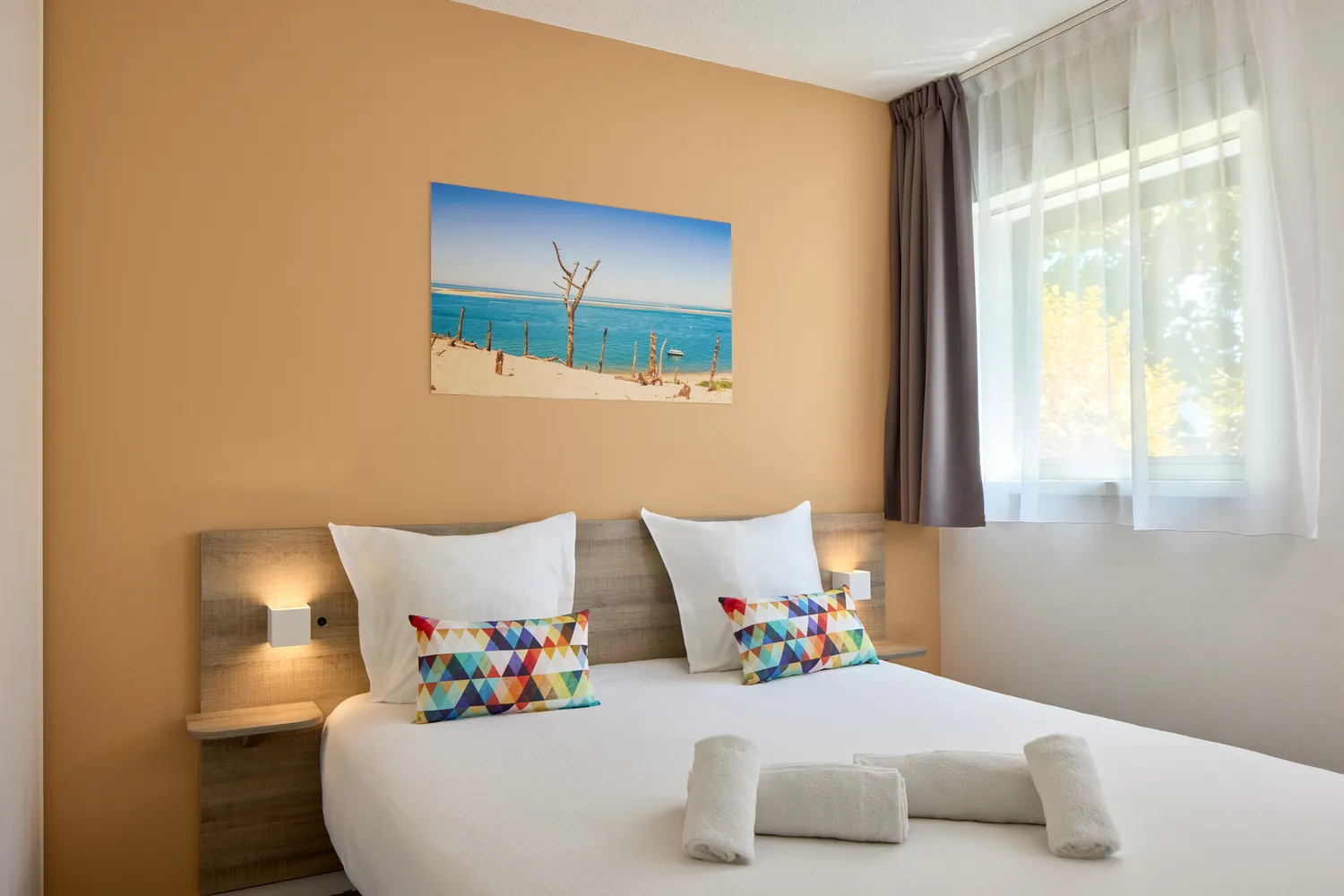 Modern bedroom with double bed, light beige walls, beach landscape artwork above the headboard, colorful geometric pillows, two rolled white towels, integrated bedside lamps, window with dual curtains overlooking greenery.