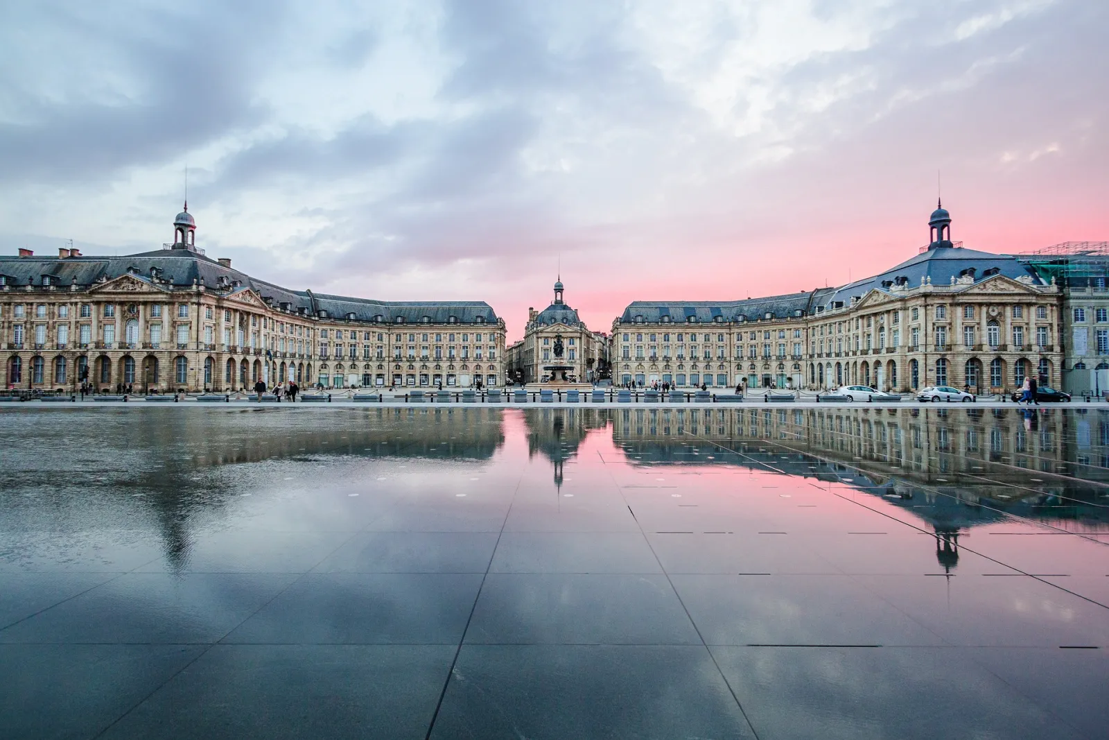 Bordeaux's Place de la Bourse at sunset, featuring symmetrical 18th-century classical buildings reflected in the water mirror, beneath a pink and cloudy sky.
