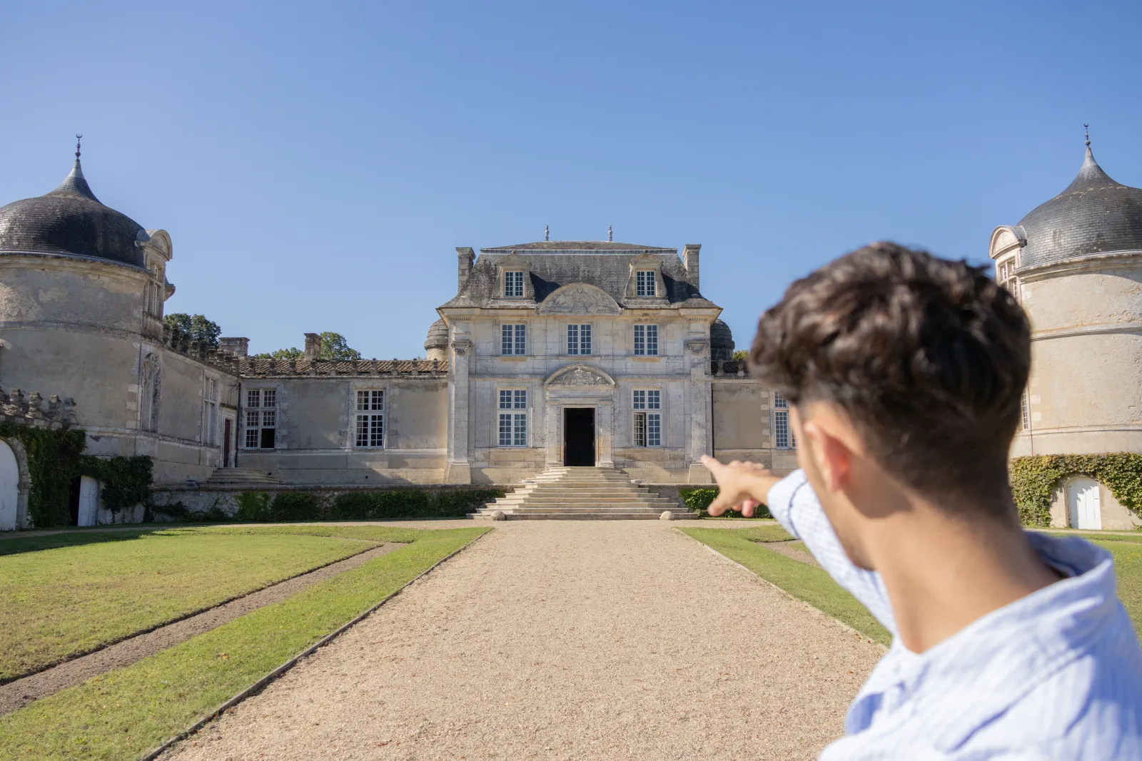 A visitor gazing at Château de Malle, a classical French estate with domed pavilions in Bordeaux, surrounded by manicured lawns and a gravel courtyard.