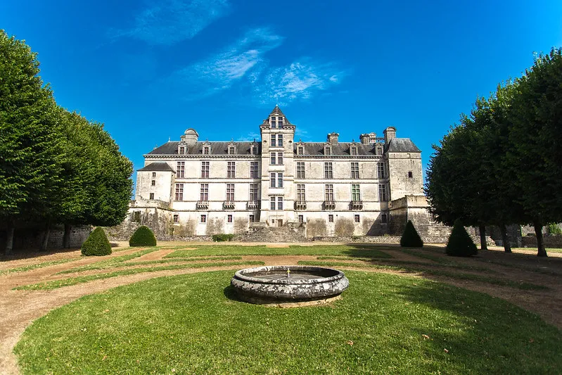 Front view of Cadillac Castle featuring its Renaissance facade, framed by manicured trees and a circular basin in the formal French gardens.