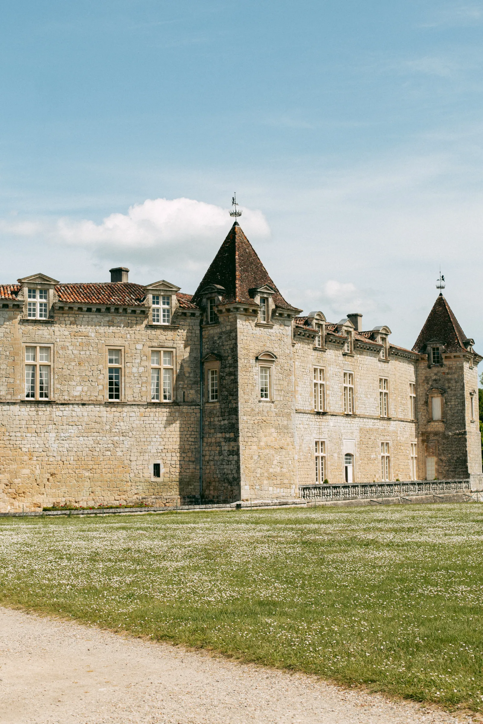 Royal Castle of Cazeneuve, medieval stone fortress with conical ivy-covered towers, surrounded by green lawn and gravel pathway, under a clear blue sky.
