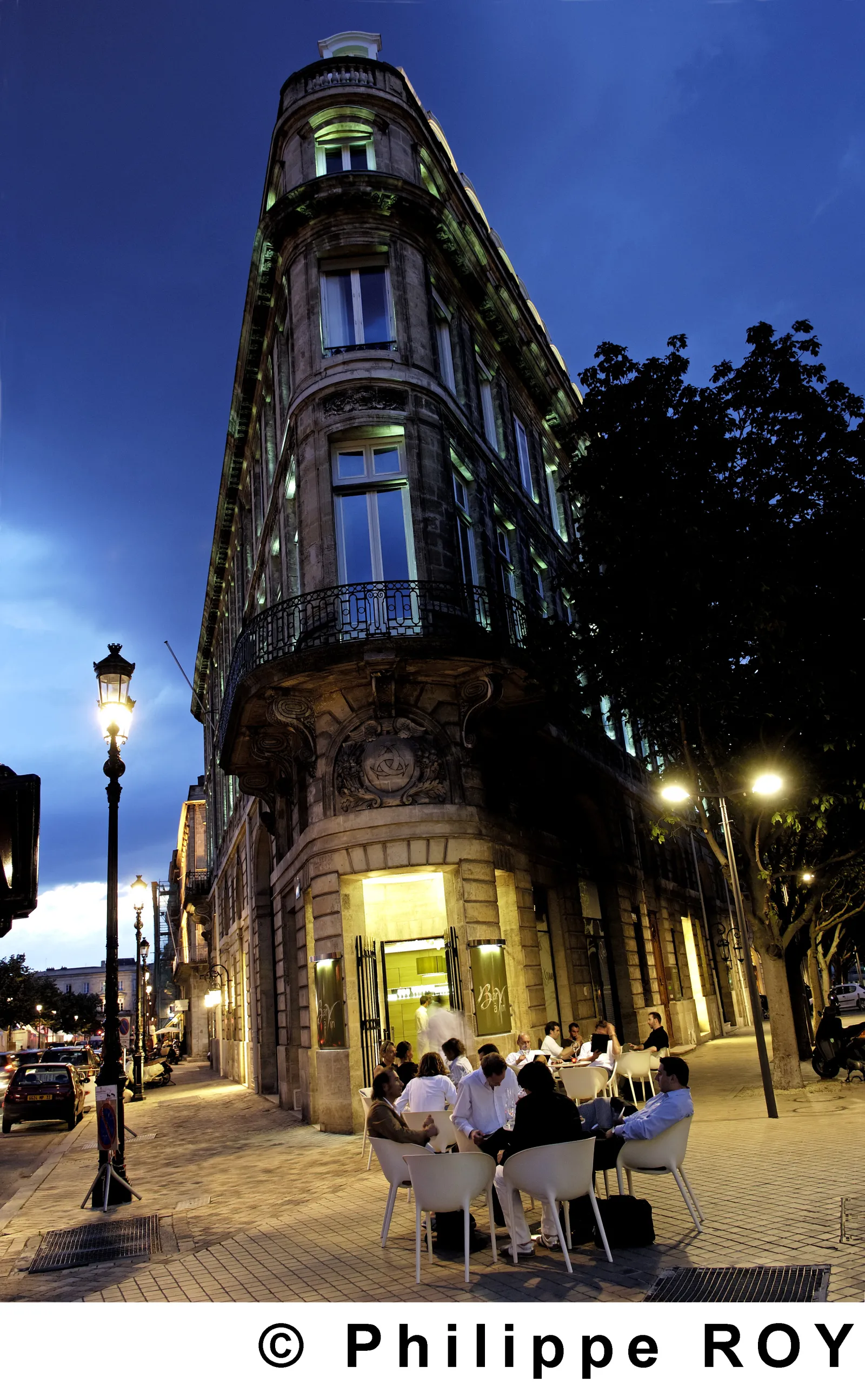 Illuminated facade of the CIVB Wine Bar in Bordeaux, Haussmann-style building with corner turret, outdoor terrace with guests at dusk, vintage street lamp and trees in background