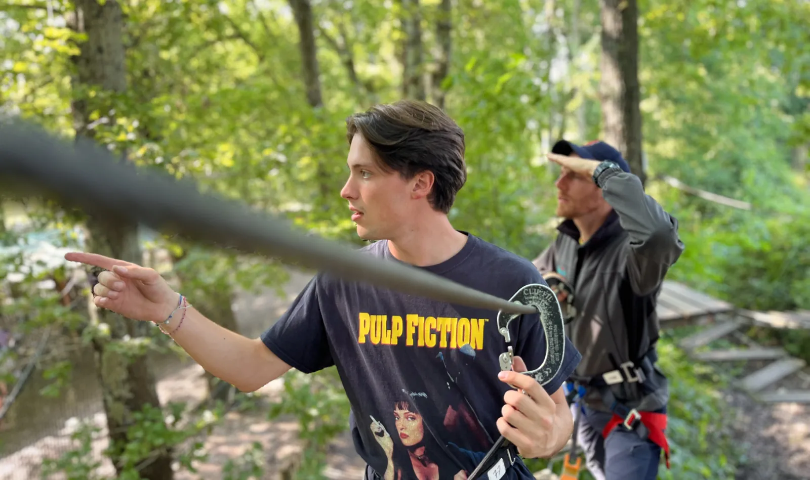 A young man wearing a Pulp Fiction t-shirt points ahead in a wooded area while participating in a treetop adventure park activity, with another participant visible in the background.