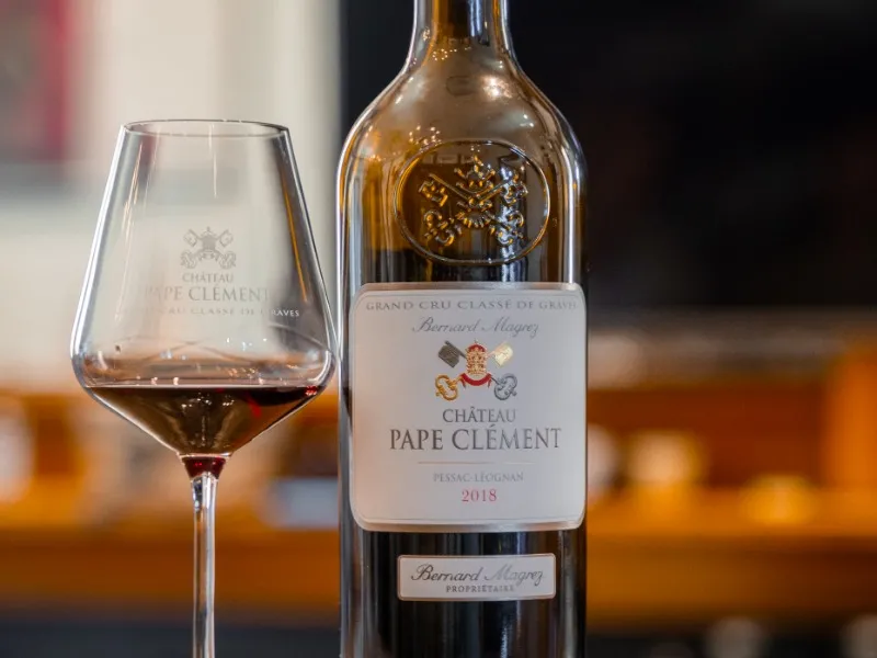 Bottle of Château Pape Clément 2018, Grand Cru Classé de Graves, with a glass of red wine against a blurred golden background.