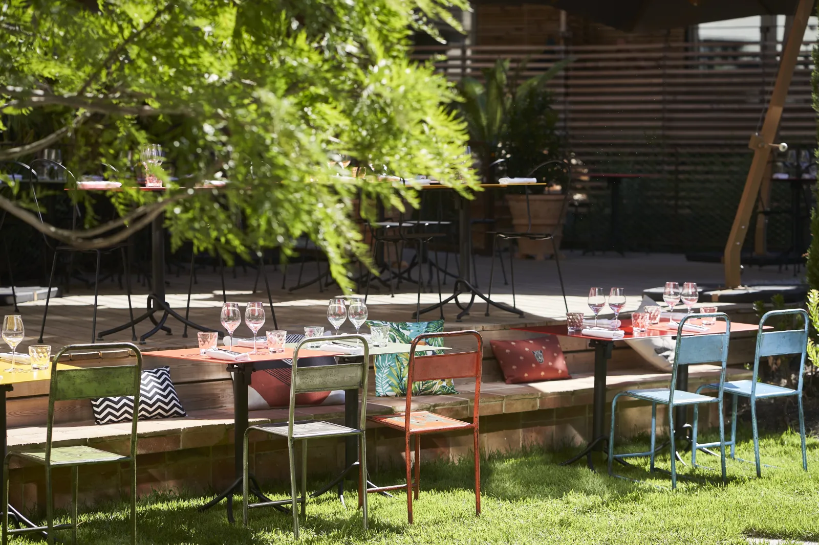 Outdoor terrace of Hôtel La Zoologie in Bordeaux featuring wooden tables, colorful metal chairs, table settings, wine glasses and lush hanging greenery providing natural shade.