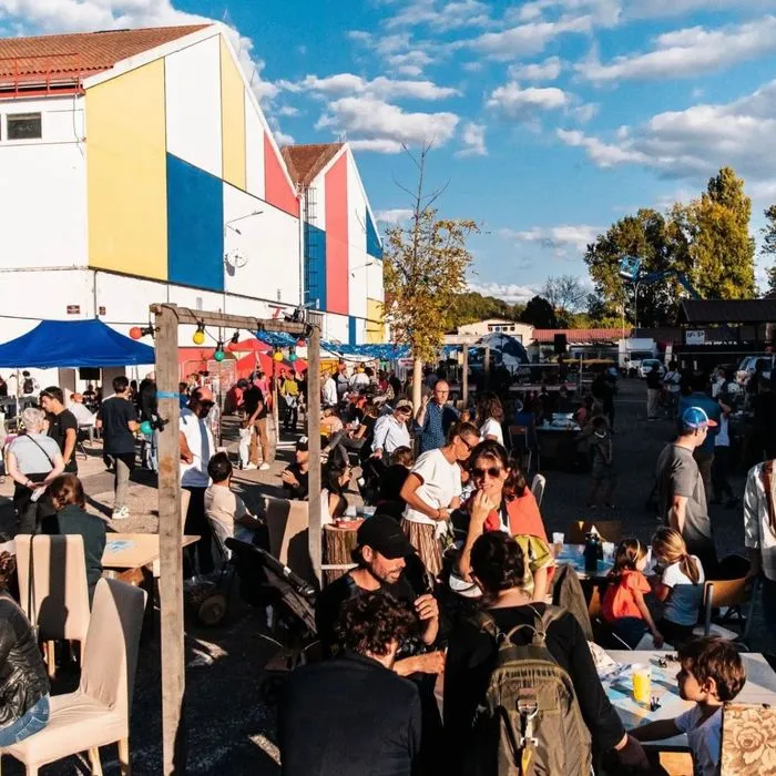 Festive crowd at the 4th edition of La Cité Bleue en fête in Bordeaux, with a colorful building featuring yellow, blue and red facades, tents, visitors and food trucks under clear blue skies.