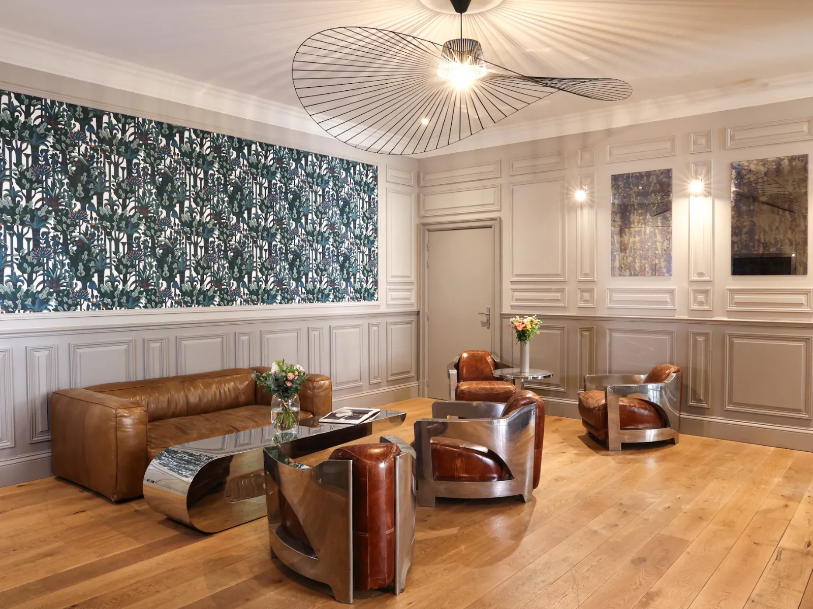 Elegant lounge of a boutique hotel featuring a cognac leather sofa, modern armchairs, stainless steel designer furniture, dark blue botanical patterned wallpaper, white wall paneling and contemporary ceiling fan.