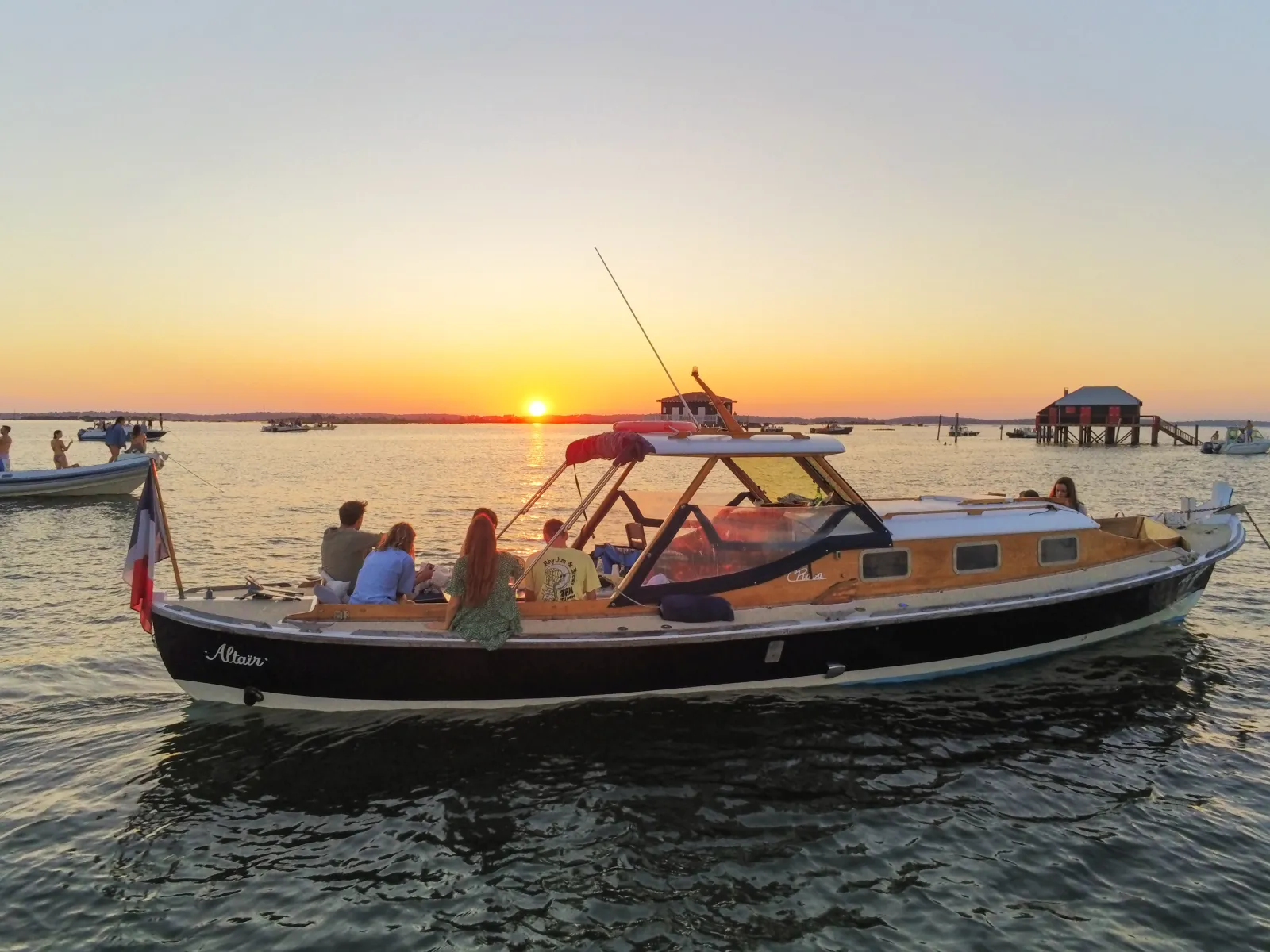 The Altaïr pinasse, a traditional orange and black Bordeaux boat, carries passengers during sunset on the Gironde estuary, with other vessels and oyster huts visible in the background.