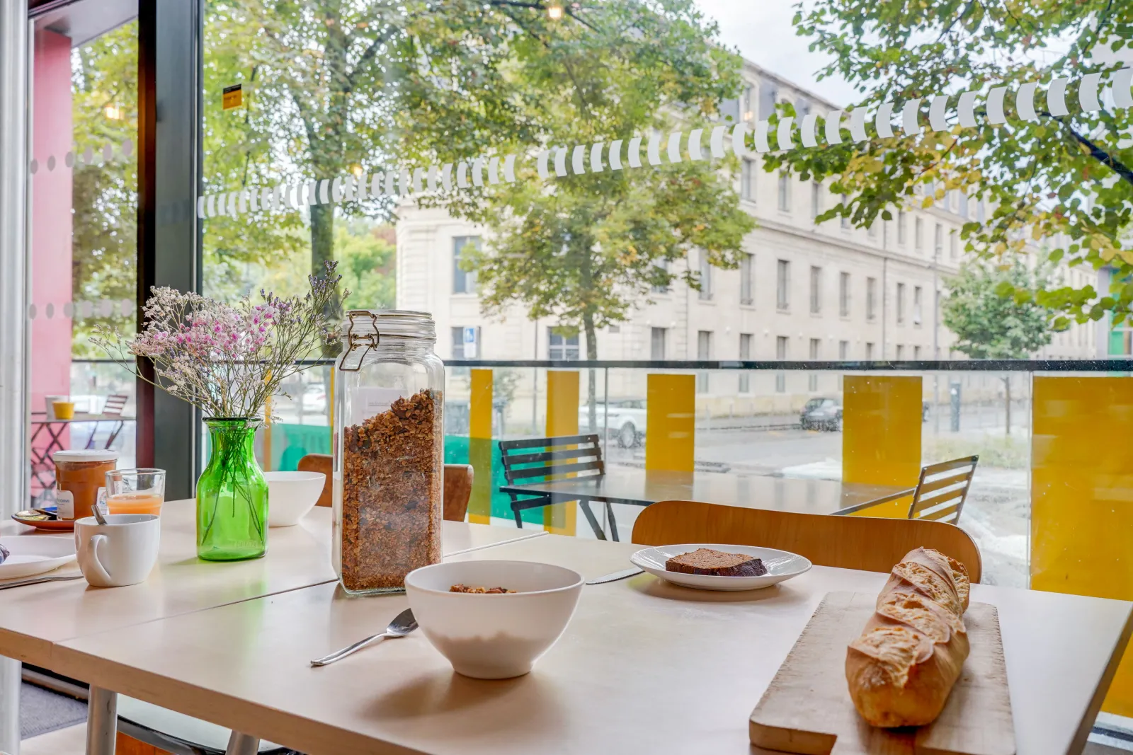 Bright dining room of Bordeaux Youth Hostel featuring a white table with breakfast spread, green flower vase, and panoramic view of the courtyard with Haussmann-style buildings.
