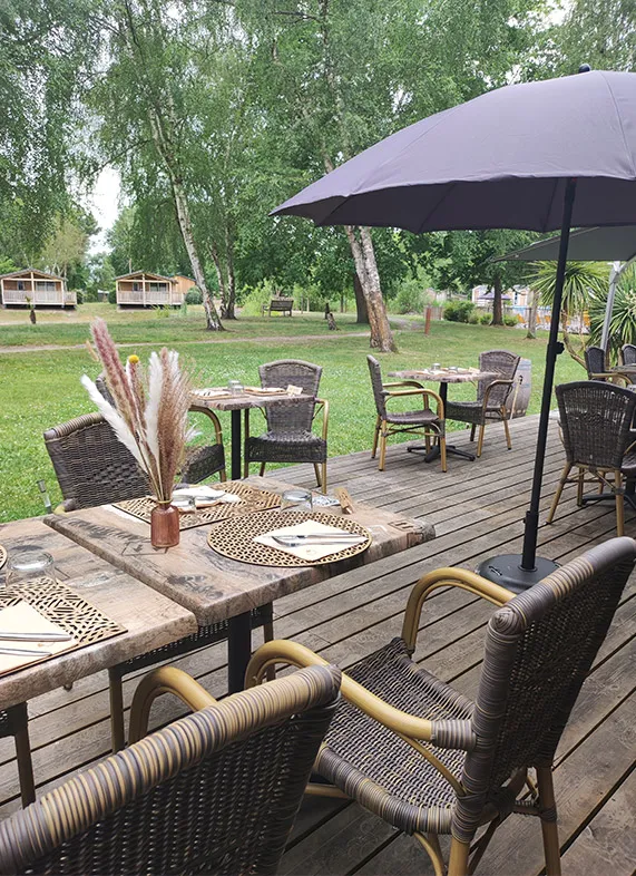 Outdoor dining terrace at Bistrot du Lac in Bordeaux featuring set tables, wicker chairs, black umbrella, floral decorations and a view of a green park with trees and small cottages in the background