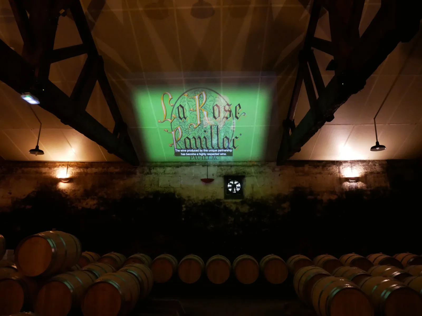 La Rose Pauillac wine cellar in Bordeaux with rows of oak barrels and illuminated projection screen displaying the brand logo
