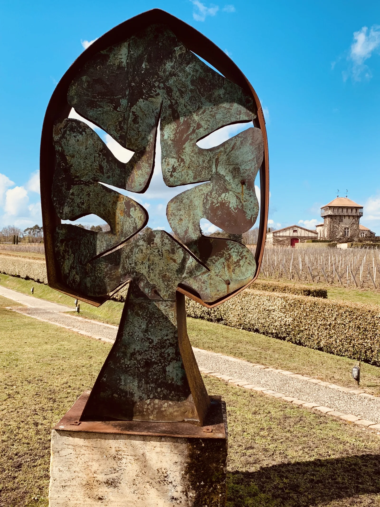 Abstract metal sculpture depicting a stylized face with weathered rust patina, positioned in the vineyards of Château Smith Haut Lafitte near Bordeaux, with winery buildings visible in the background.