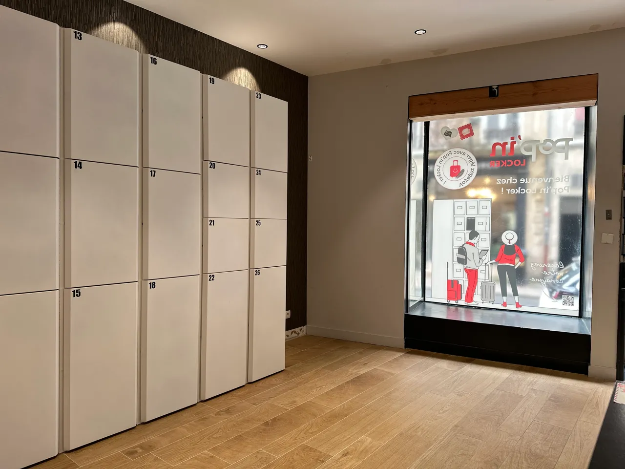 Interior of a Popin Locker automated locker service in Bordeaux, featuring a row of numbered cream and beige lockers on the left side and an illuminated display window on the right showing products and usage instructions.