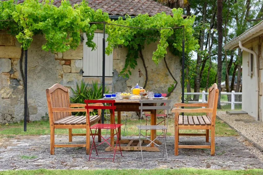 Outdoor patio at Bastide de Siran in Bordeaux featuring a wooden table, teak lounge chairs, pergola draped with lush green vines, rustic stone building and countryside setting.