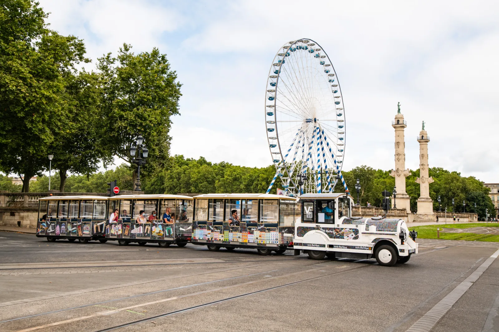 White electric tourist train in Bordeaux passing by the Big Wheel and the Concorde Square columns