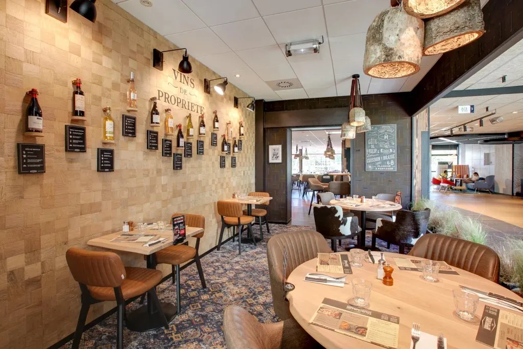 Interior of Hippopotamus restaurant in Bordeaux featuring round and rectangular dining tables, brown leather chairs, stone wall with proprietary wines display, glass pendant lights, and view of adjacent dining area