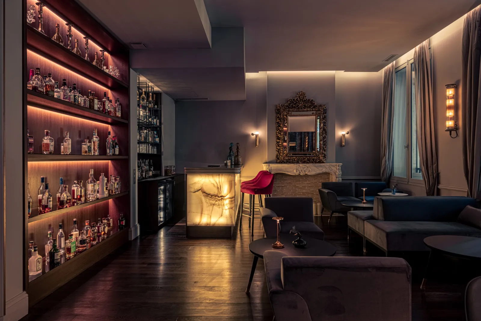Elegant bar at Hôtel Villas Foch in Bordeaux featuring illuminated shelves with spirits bottles, luminous counter, dark furniture, ornate mirror and warm ambient lighting