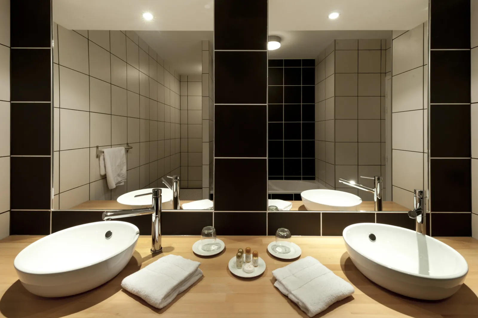 Modern bathroom at Château La Chèze in Bordeaux featuring two white oval sinks, large mirror, black and beige tile, wooden countertop, and luxury amenities