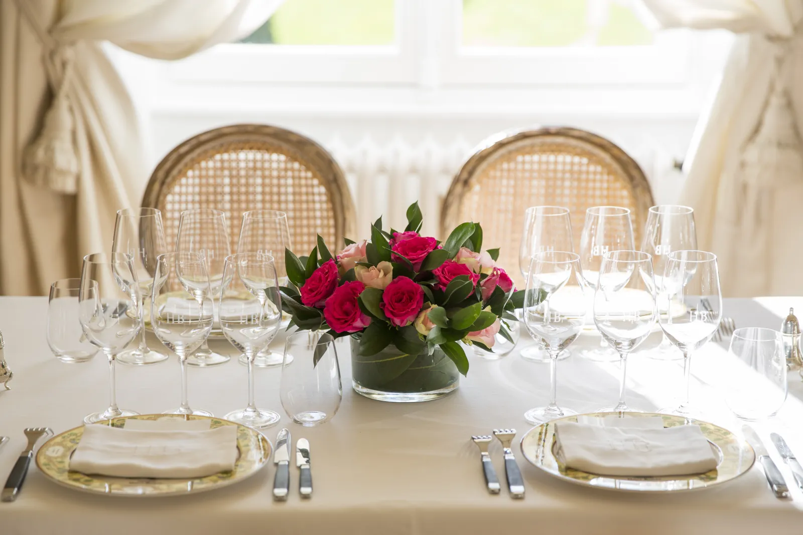 Elegant table setting at Château Haut-Bailly in Bordeaux featuring gold-rimmed plates, crystal glassware, white napkins, and a central arrangement of red roses with rattan chairs in the background.