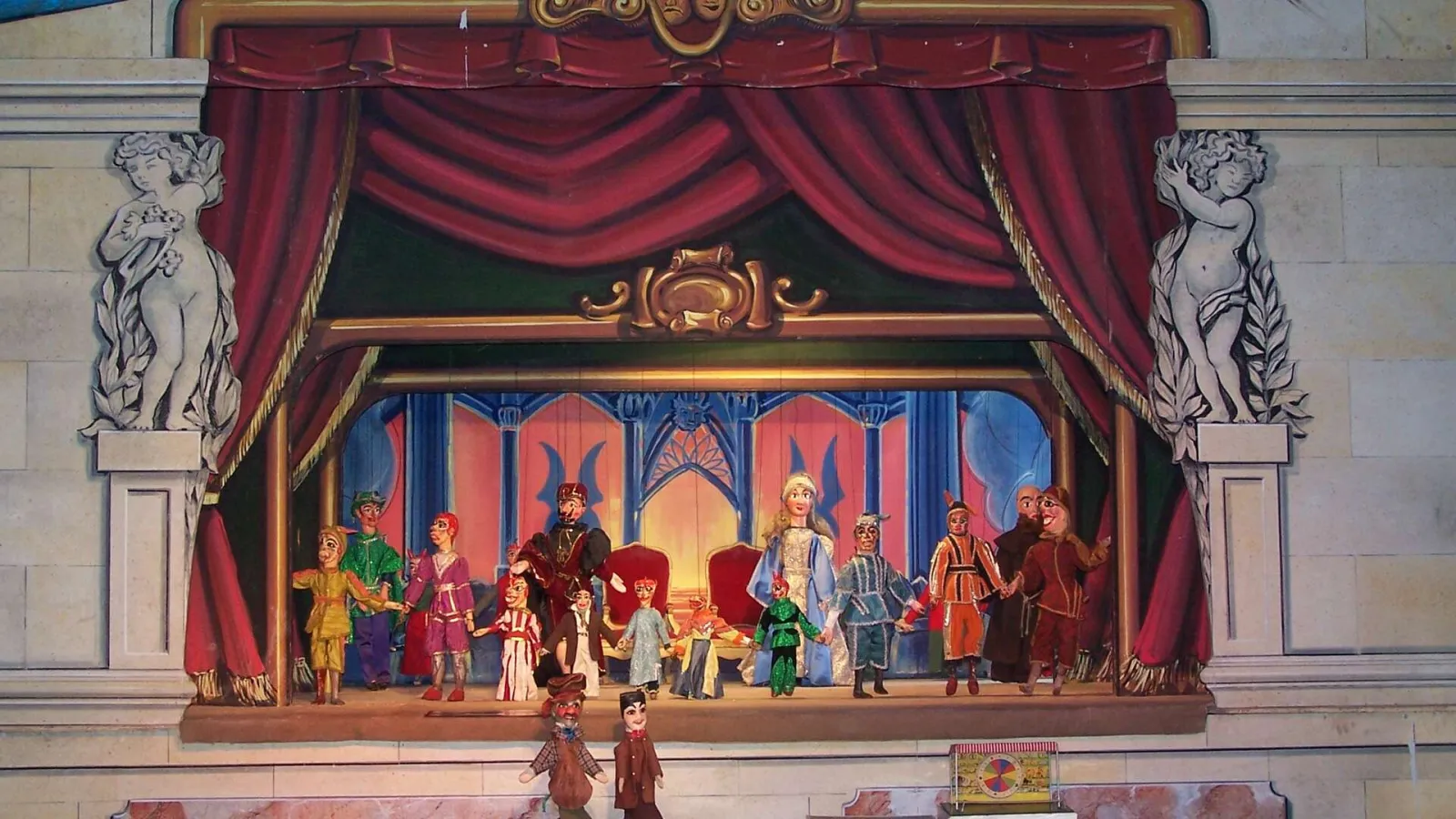 Puppet theater stage at Guignol Guérin in Bordeaux, depicting a royal court scene with characters in period costumes on a platform decorated with red curtains and golden ornaments.