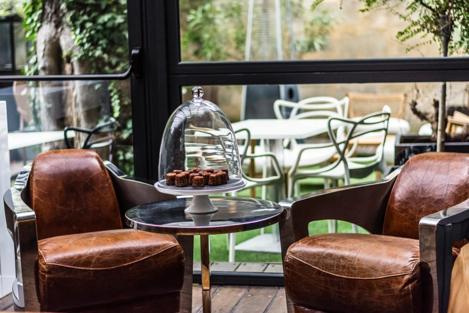 Lounge area of a Bordeaux boutique hotel featuring two brown leather armchairs facing a black side table with a glass dome cake cover, overlooking a terrace with green chairs