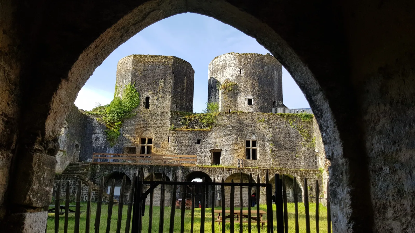 View of Villandraut Castle near Bordeaux through a stone archway, featuring two cylindrical towers in the background, fortified walls with vegetation, and an iron gate in the foreground.