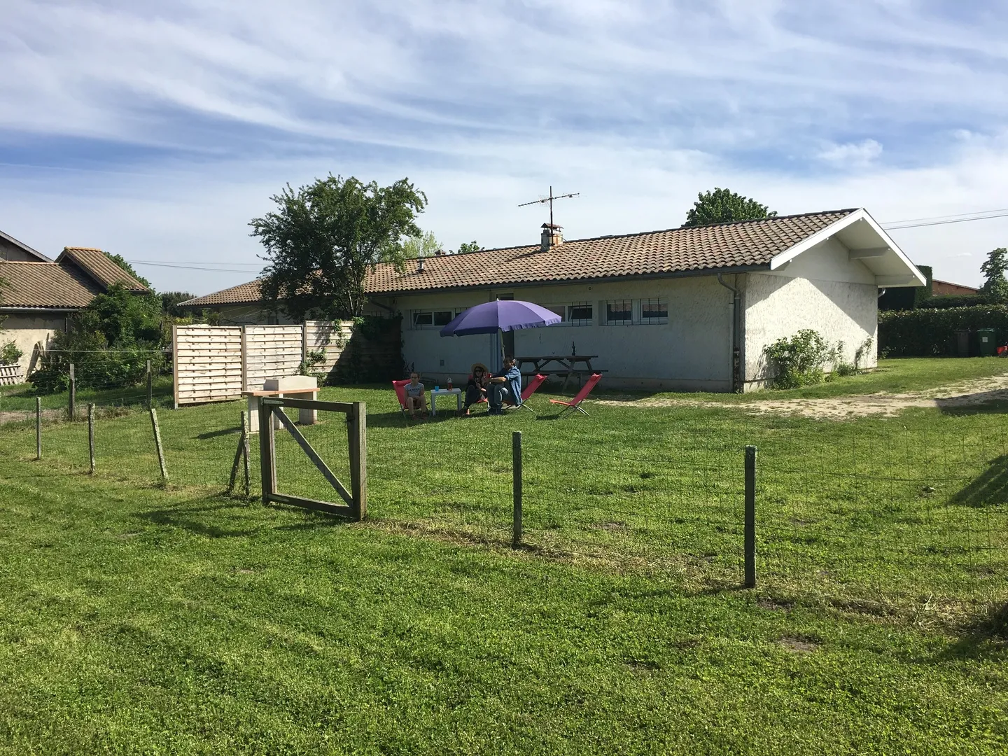 Exterior view of a rural cottage with a family under an umbrella in a fenced garden, in the Bordeaux region