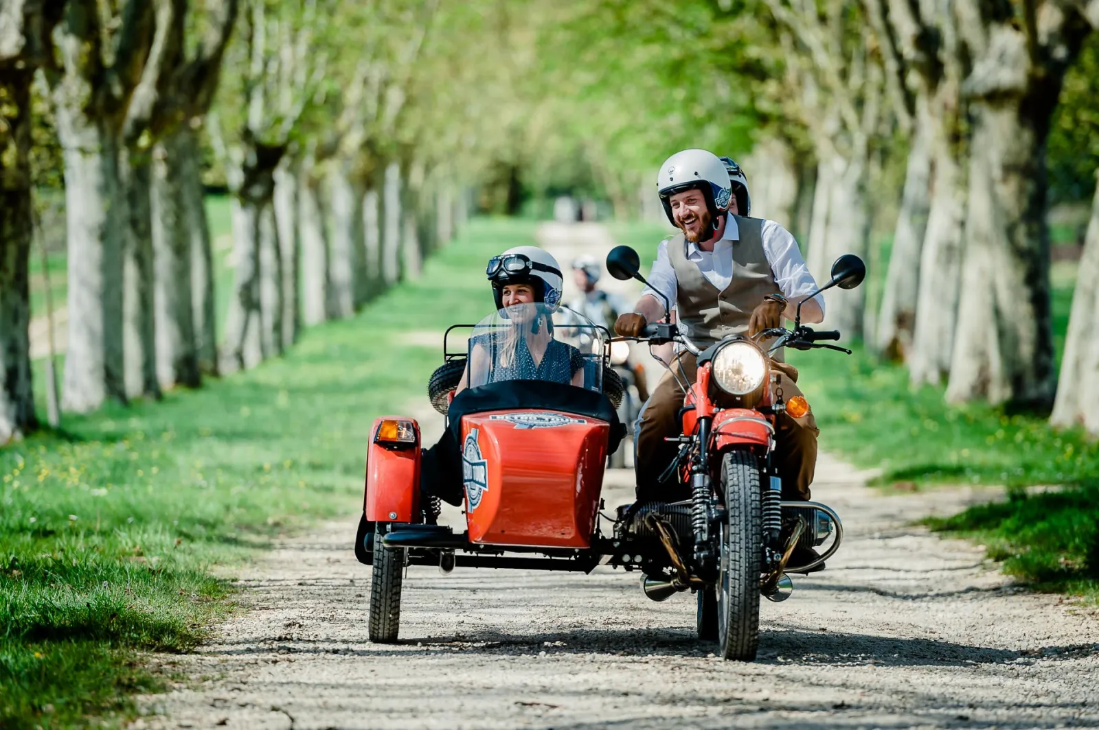 Two helmeted riders on a vintage motorcycle with red sidecar driving through a tree-lined avenue in Bordeaux during a retro tour.