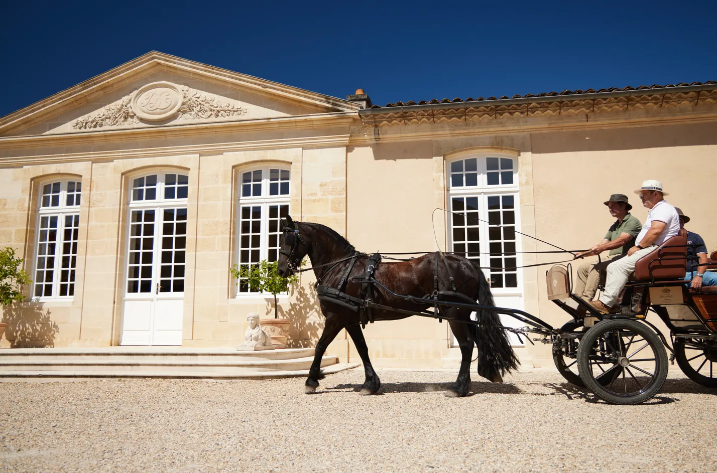 A black horse-drawn cart with three passengers in front of the classical facade of Château Desmirail in Bordeaux, under clear blue sky