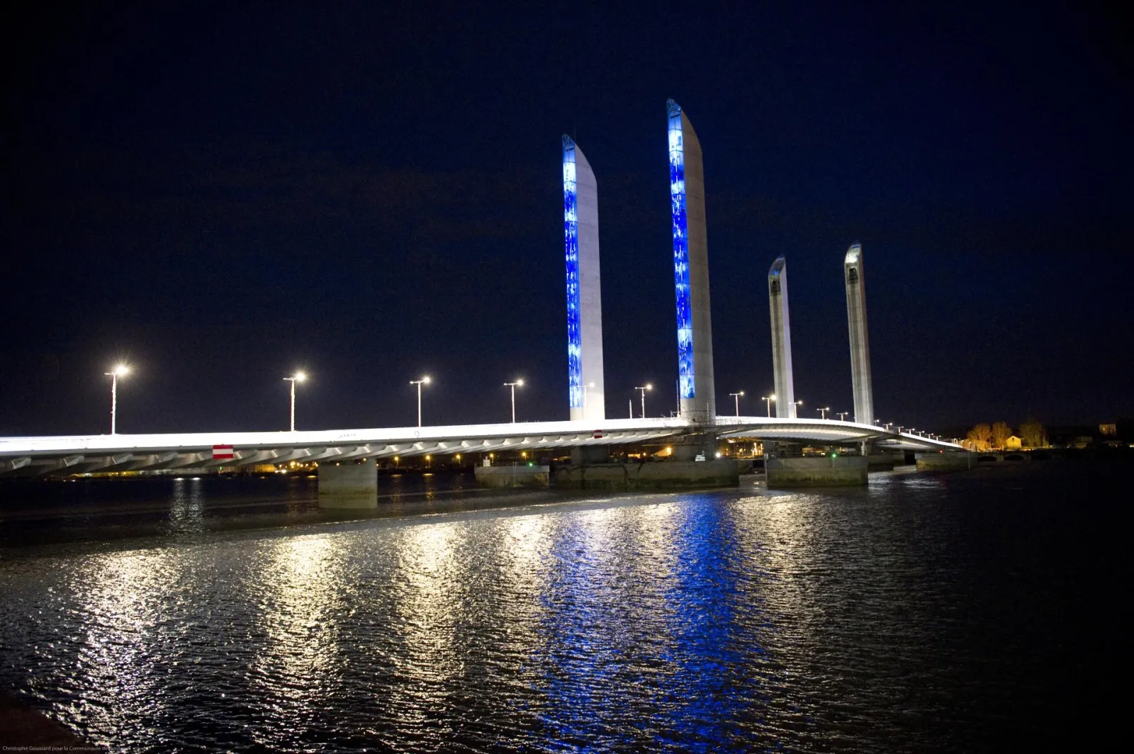 Bordeaux's Jacques Chaban Delmas Bridge lit up at night, featuring four blue illuminated vertical pylons reflected in the Garonne River