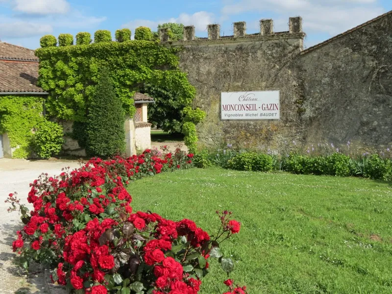 Entrance to Château Montsegur-Gazin in Bordeaux, featuring stone walls with crenellations, red roses in the foreground, and Michel Baudet wine estate sign.
