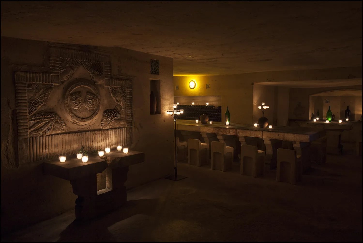 Underground cellars of Château de La Rivière in Bordeaux, lit by white candles, featuring sculpted wall reliefs, chairs, and wine bottles displayed on shelves
