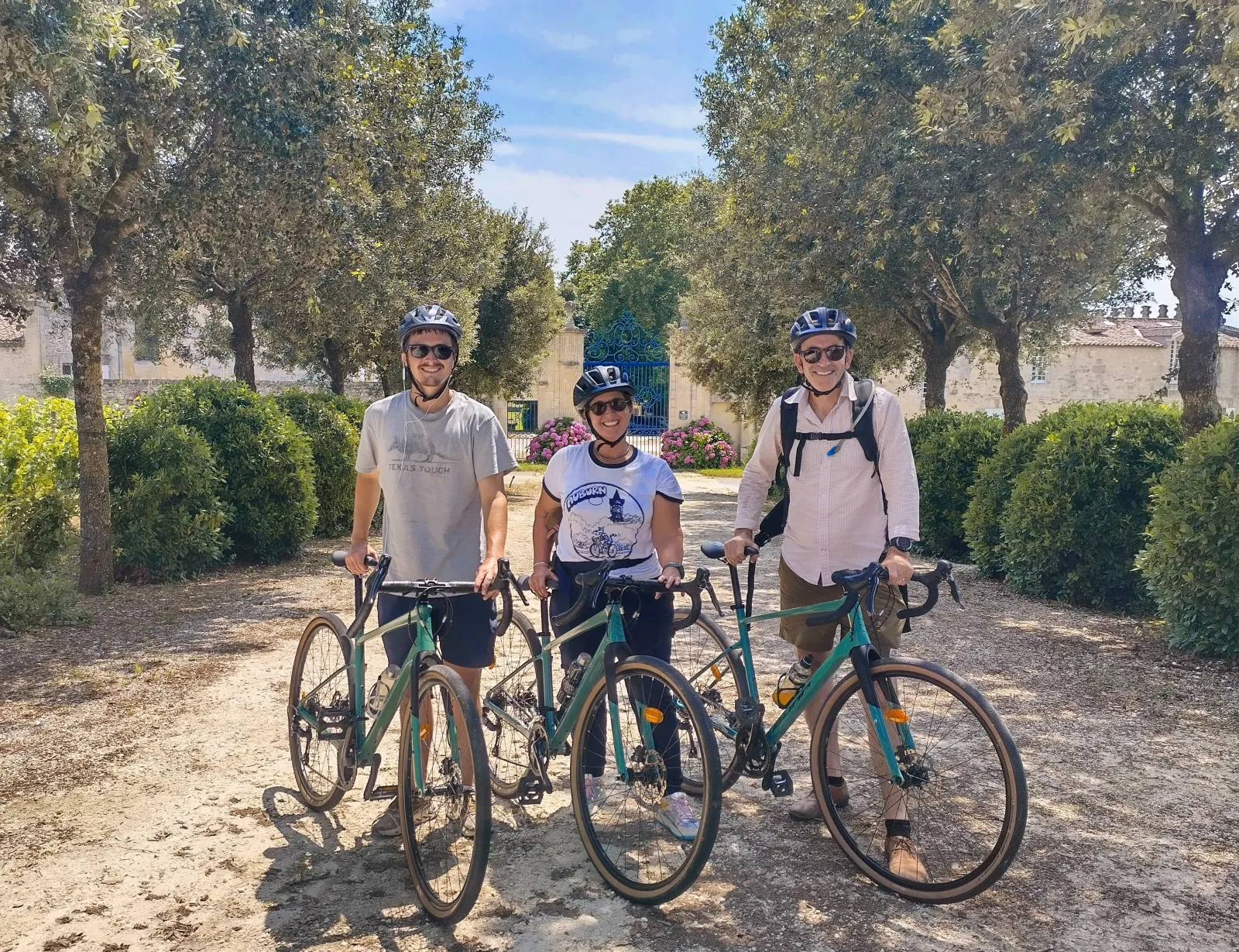 Three cyclists wearing helmets with green gravel bikes pose on a tree-lined path in Bordeaux vineyards