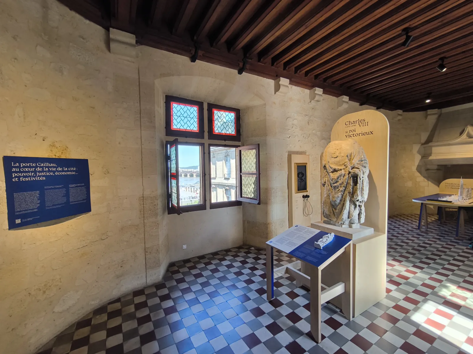 Interior of the Porte Cailhau museum in Bordeaux featuring historical exhibits with informational panels, a statue of Charles VIII, and a view of the Garonne River through latticed windows.
