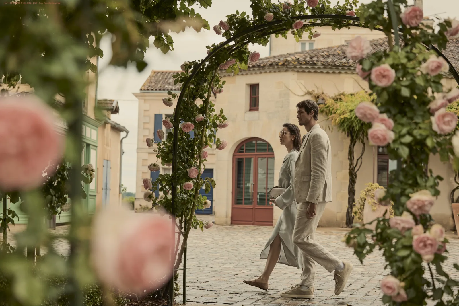 A couple in beige linen clothing strolls through a paved courtyard surrounded by pink climbing roses and Mediterranean-style stone buildings.