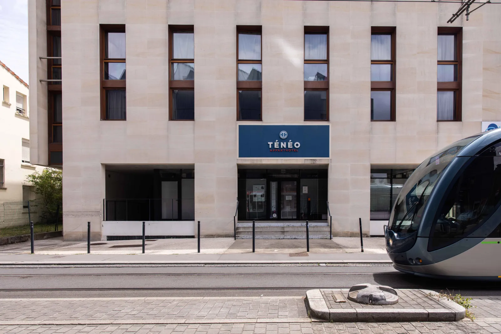 Front view of Ténéo Bordeaux-Maritime hotel featuring a modern stone façade, ground floor entrance, regular window rows, and a tram passing by the building.