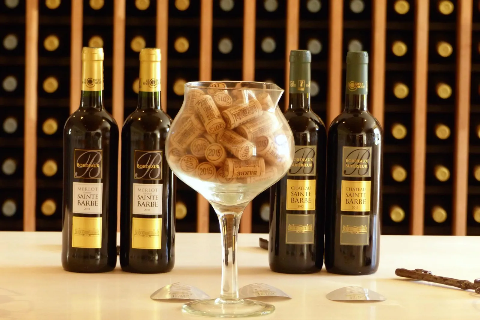 Five bottles of Bordeaux Château Sainte Barbe wine arranged around a glass filled with corks, in front of a gold and black-toned wine cellar wall.
