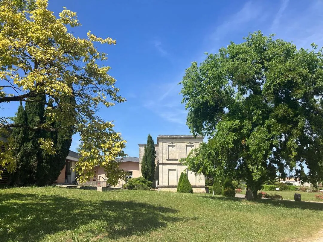 View of Château Luchey-Halde in Bordeaux featuring stone buildings surrounded by century-old trees and a green lawn under a blue sky.