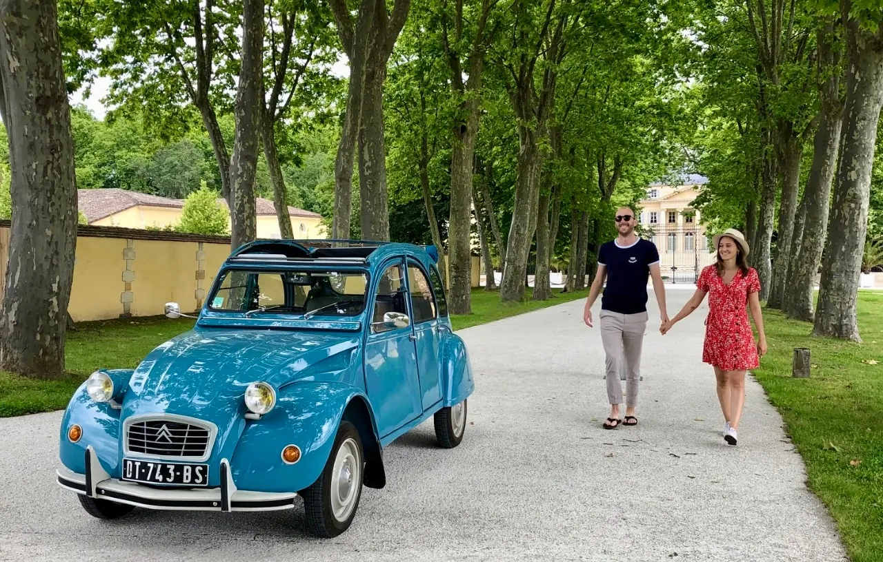 A turquoise Citroën 2CV with an open roof parked under a tree-lined avenue in Bordeaux, with a couple walking nearby and historic buildings in the background.