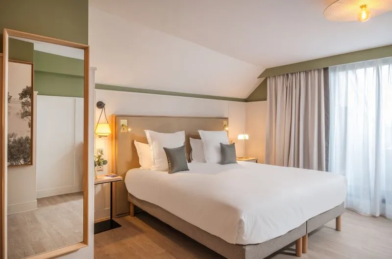 Modern hotel bedroom featuring a white double bed, green and white walls, contemporary furnishings, large window with curtains, and attached bathroom.