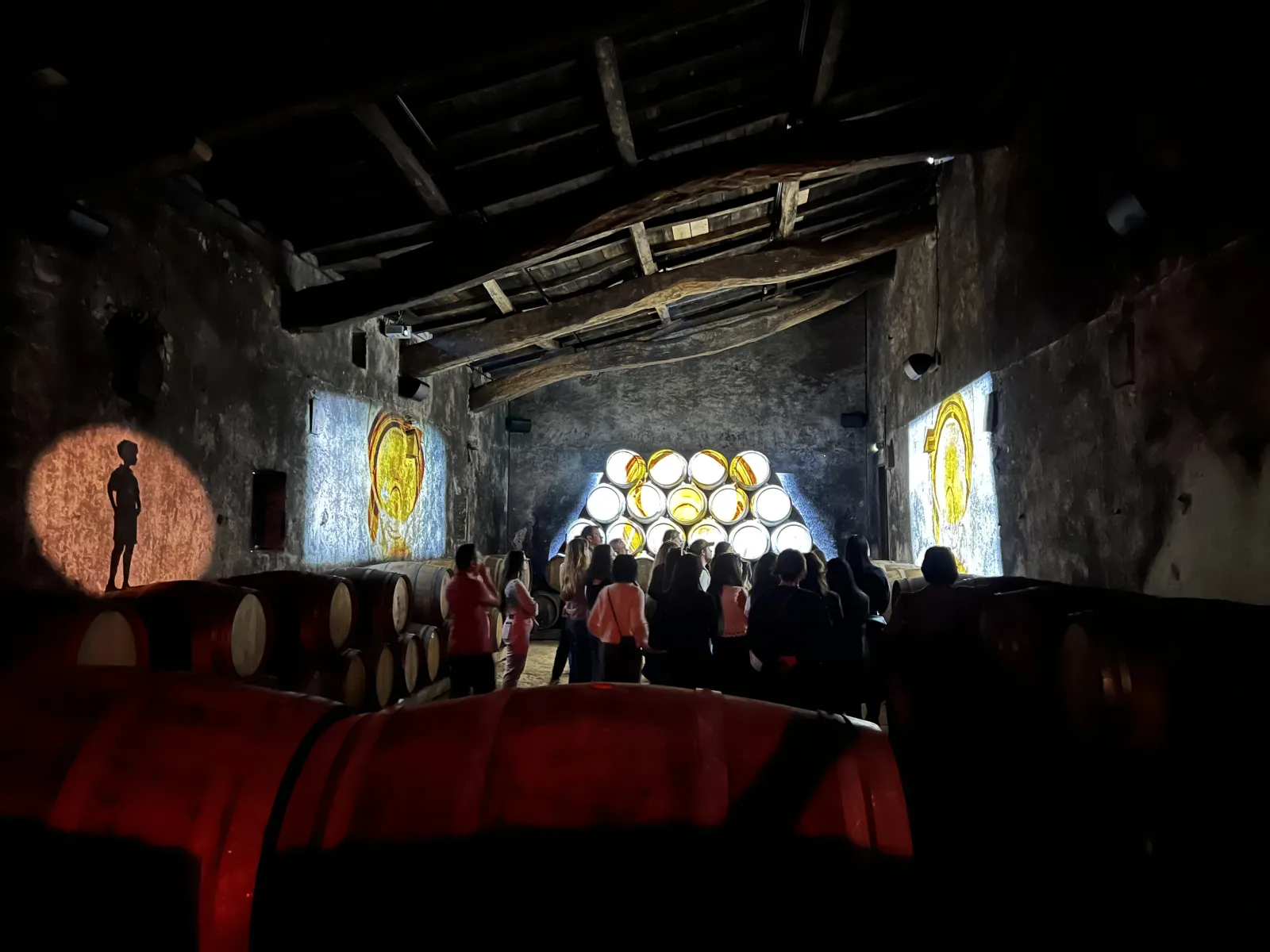 Immersive art installation in a Bordeaux castle cellar featuring golden and red light projections, wine barrels, and visitors experiencing the exhibition.