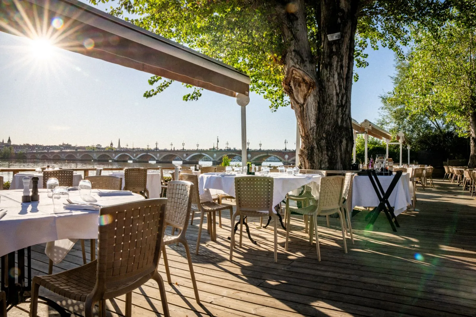 Sunny waterfront terrace at Café du Port in Bordeaux with set tables, wicker chairs, wooden pergola, view of the Stone Bridge and Garonne river