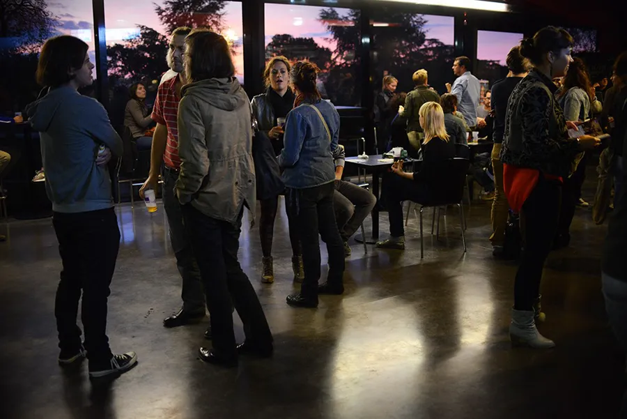 Group of people standing and chatting in a modern hall at Rocher de Palmer in Cenon, with large windows overlooking a sunset.