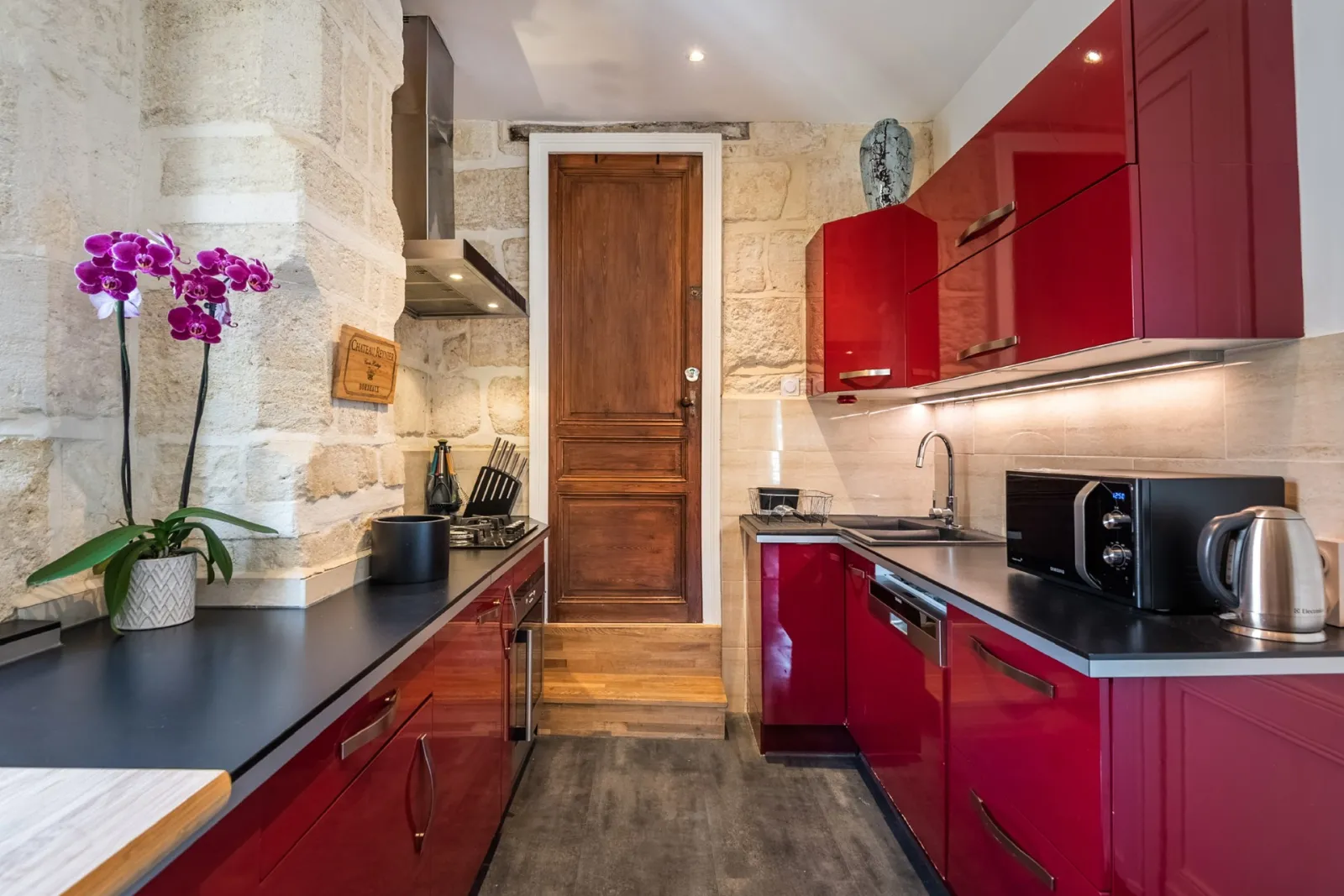Modern kitchen featuring bright red cabinetry, exposed beige stone walls, black countertops, stainless steel appliances and purple orchid on the work surface in Bordeaux La Monnoye
