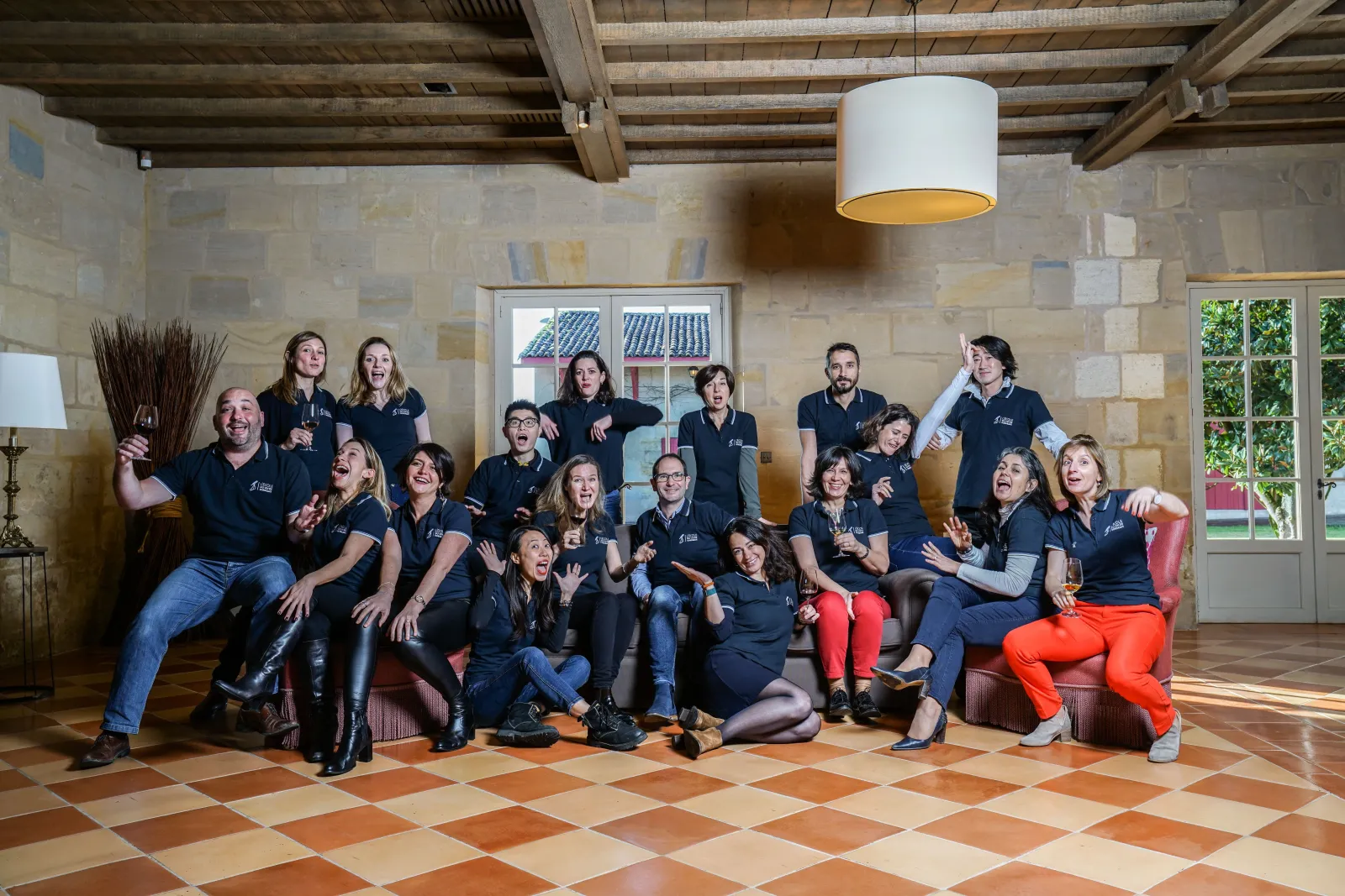Group of approximately 25 people wearing navy blue polo shirts posing together in a modern room at the Bordeaux Wine School, featuring exposed wooden ceiling and checkered tile flooring.