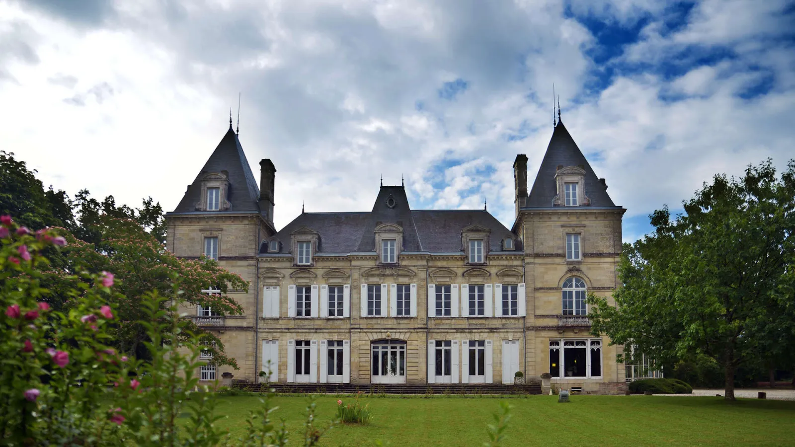 Front facade of the National Health Insurance Museum in Bordeaux, a Second Empire-style château with two pointed towers, surrounded by green lawns and pink flowers.