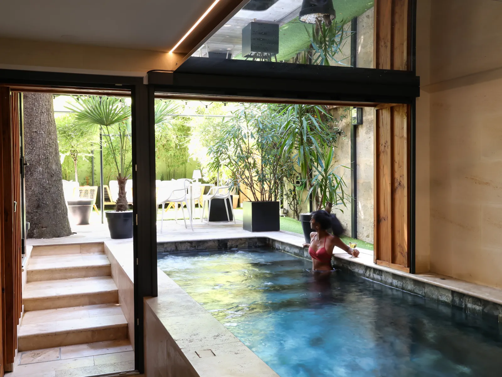 A woman relaxes in a modern indoor pool at a luxury spa boutique in Bordeaux, overlooking a lush garden through large glass doors.