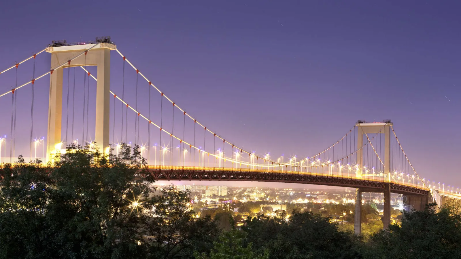The Aquitaine Bridge in Bordeaux lit up at night, featuring its golden suspended cables and two massive towers overlooking the Garonne river and the illuminated city beyond.