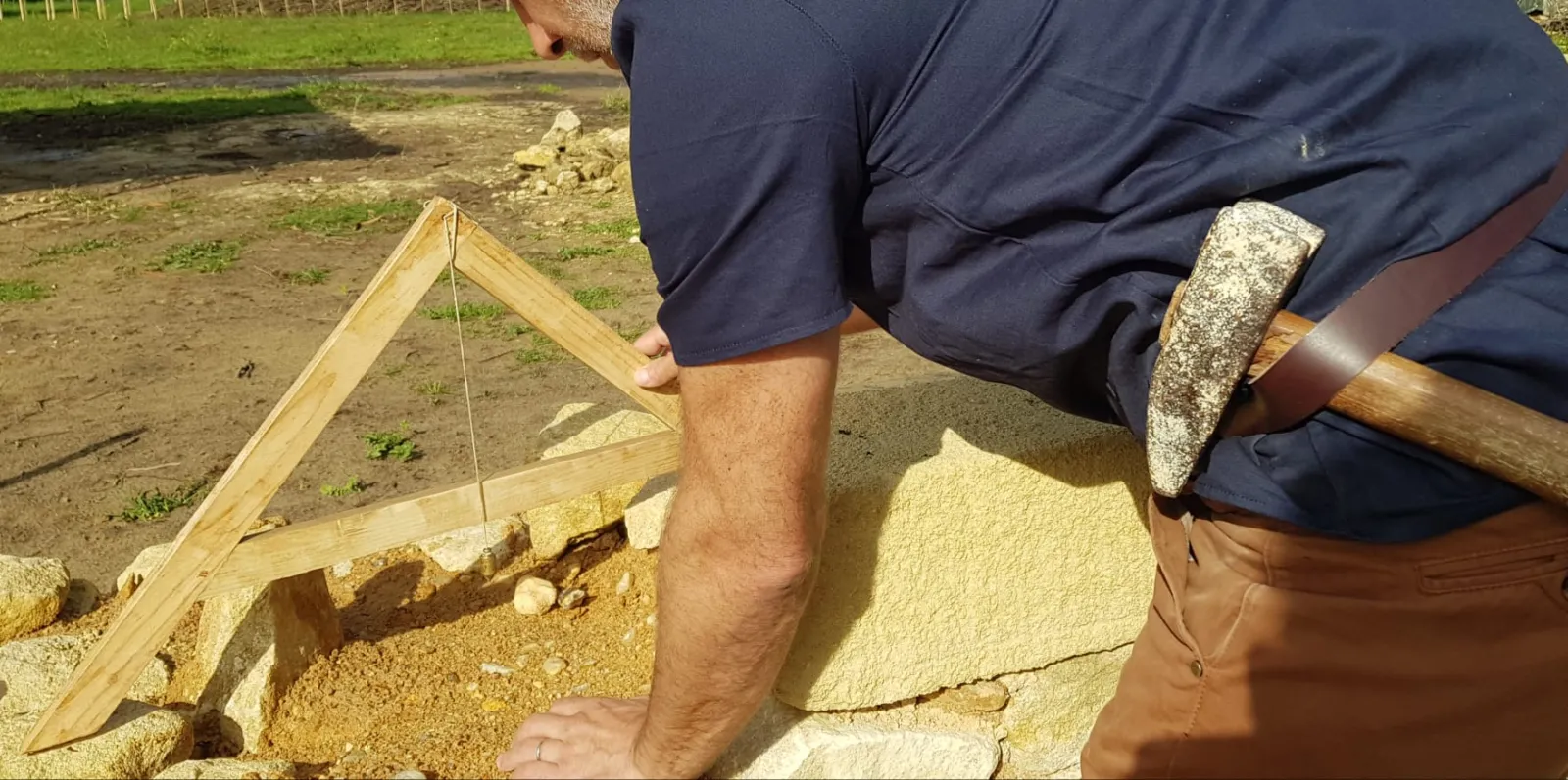 Stonemason chiseling a stone block by hand with a chisel and hammer on a medieval reconstruction site in Bordeaux, with construction tools and materials visible.